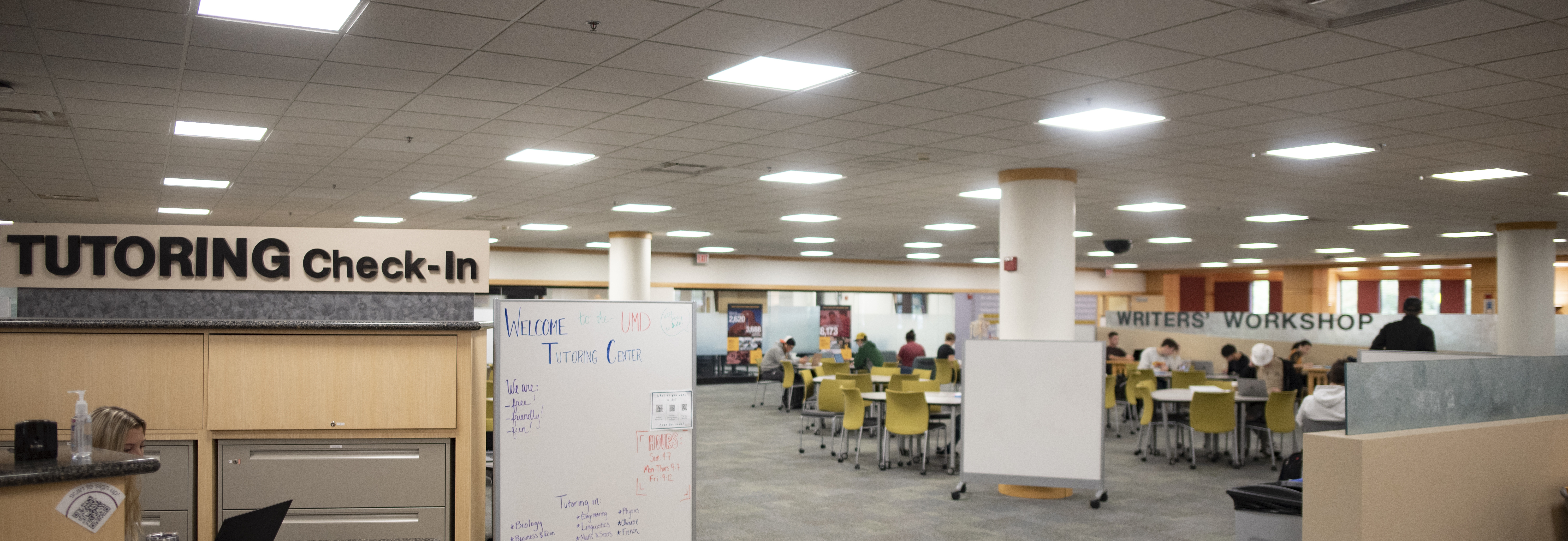 photo of an brightly-lit space full of tables in the library, tutoring center on one side, writers' workshop on the other